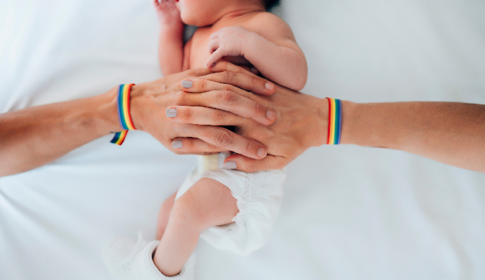 Due mani con un braccialetto arcobaleno gay si tengono per mano e abbracciano il loro figlio neonato.
